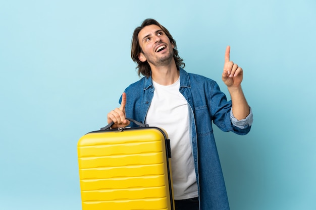 Jonge knappe man geïsoleerd op blauwe muur in vakantie met reiskoffer en omhoog