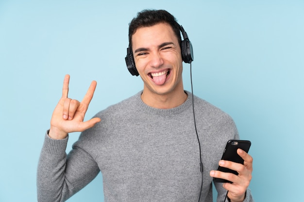 Jonge knappe man geïsoleerd op blauwe muur luisteren muziek met een mobiele rock gebaar maken