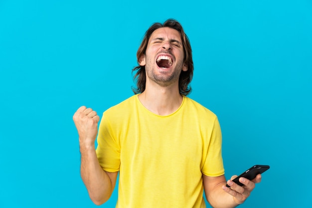 Jonge knappe man geïsoleerd op blauwe muur met behulp van mobiele telefoon en overwinning gebaar doen