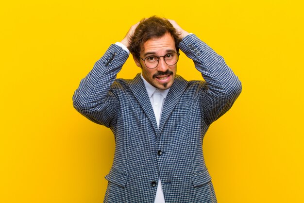 Jonge knappe man gevoel gestrest, bezorgd, angstig of bang, met handen op het hoofd, in paniek bij vergissing tegen oranje muur