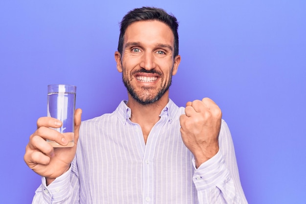 Jonge knappe man glas water drinken tot verfrissing over geïsoleerde paarse achtergrond schreeuwend trots vieren overwinning en succes erg opgewonden met opgeheven arm