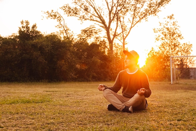 Jonge knappe man in een meditatieve positie zit op een grasveld op een achtergrond van bos en zonsondergang. Concept van vrijheid ontspanning. Plaats voor tekst of reclame