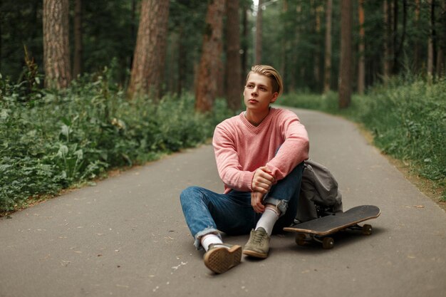 Jonge knappe man in een roze modieuze trui met een skateboard zit op asfalt in het park