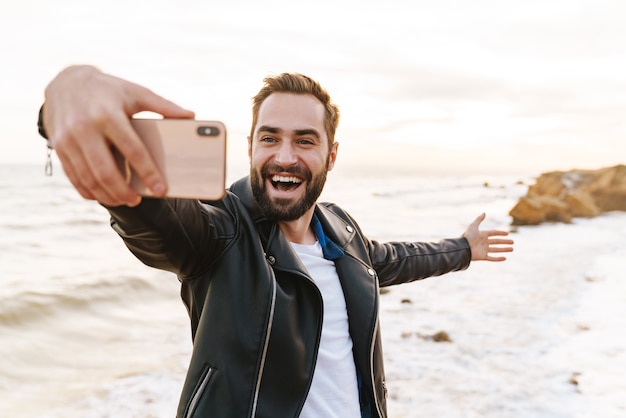 Jonge knappe man in leren jas selfie foto nemen op smartphone tijdens het wandelen langs de kust