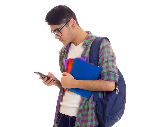 Jonge knappe man in T-shirt en geruit overhemd met rugzak die boeken vasthoudt en smartphone gebruikt