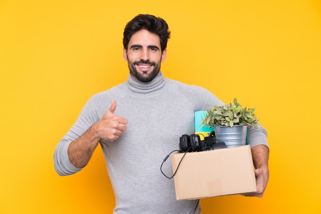 Jonge knappe man met baard een beweging maken terwijl het oppakken van een doos vol dingen over geïsoleerde muur met duimen omhoog omdat er iets goeds is gebeurd