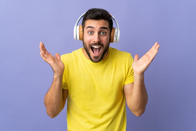 Jonge knappe man met baard geïsoleerd op paarse achtergrond verrast en muziek luisteren