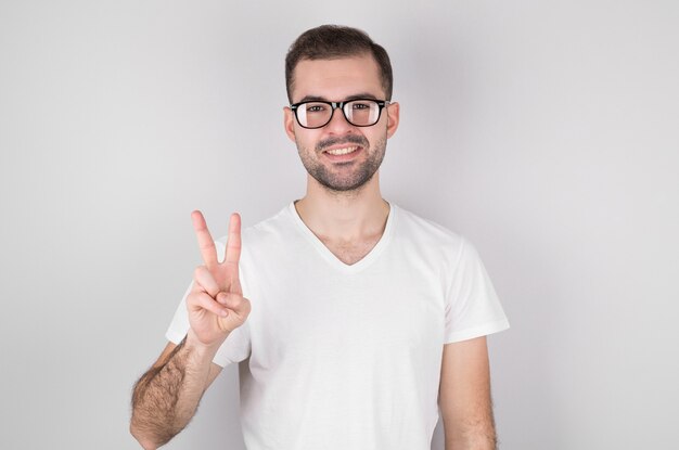 Jonge knappe man met baard, gekleed in een casual t-shirt, staande tegen een witte muur, lachend met een blij gezicht, een overwinningsteken maken.