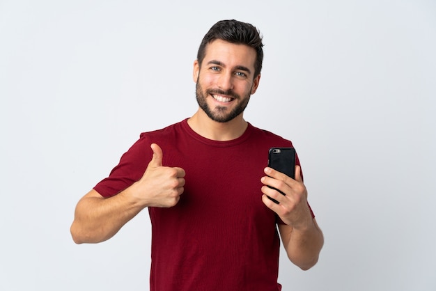 Jonge knappe man met baard met behulp van mobiele telefoon geïsoleerd op een witte muur met duimen omhoog omdat er iets goeds is gebeurd