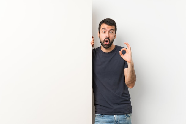 Jonge knappe man met baard met een groot leeg plakkaat verrast en het tonen van ok teken