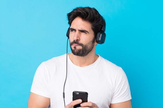 Jonge knappe man met baard over geïsoleerde blauwe muur luisteren muziek met een mobiel en denken