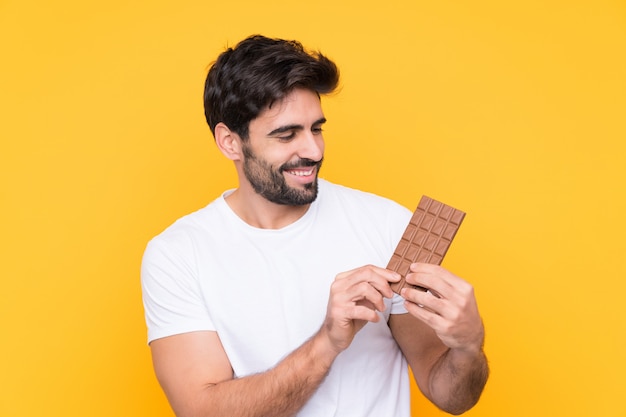 Jonge knappe man met baard over geïsoleerde gele muur die een chocoladetablet nemen en gelukkig