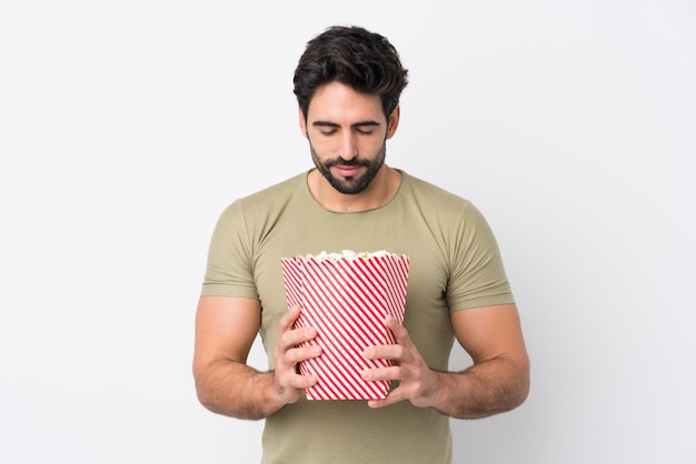 Jonge knappe man met baard over geïsoleerde witte muur met een grote emmer popcorns