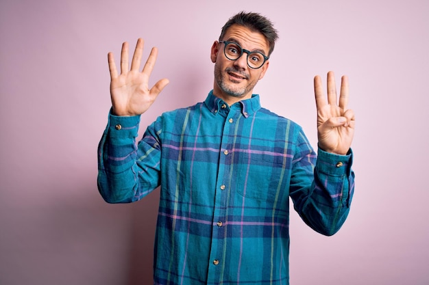 Jonge knappe man met casual shirt en bril die over een geïsoleerde roze achtergrond staat en omhoog wijst met vingers nummer acht terwijl hij zelfverzekerd en gelukkig glimlacht