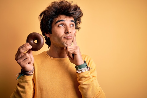 Jonge knappe man met chocoladedoughnut die over een geïsoleerde gele achtergrond staat, serieus gezicht na te denken over de vraag, erg verward idee