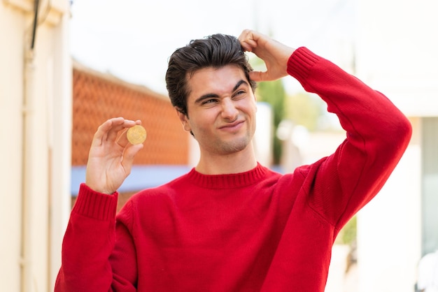 Jonge knappe man met een bitcoin buitenshuis met twijfels en een verwarde gezichtsuitdrukking