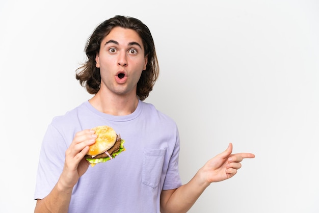Jonge knappe man met een hamburger geïsoleerd op een witte achtergrond verrast en wijzende kant