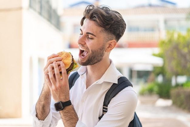 Jonge knappe man met een hamburger in de open lucht met een gelukkige uitdrukking