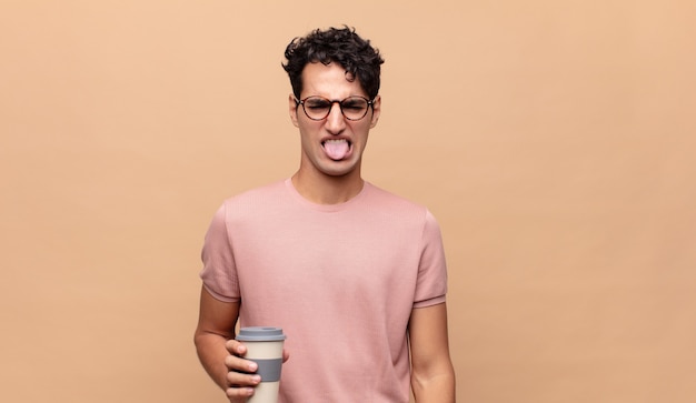 Jonge knappe man met een koffie die walgt en geïrriteerd voelt, tong uitsteekt