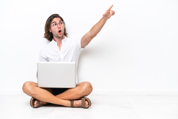 Jonge knappe man met een laptop zittend op de vloer geïsoleerd op een witte achtergrond die weg wijst