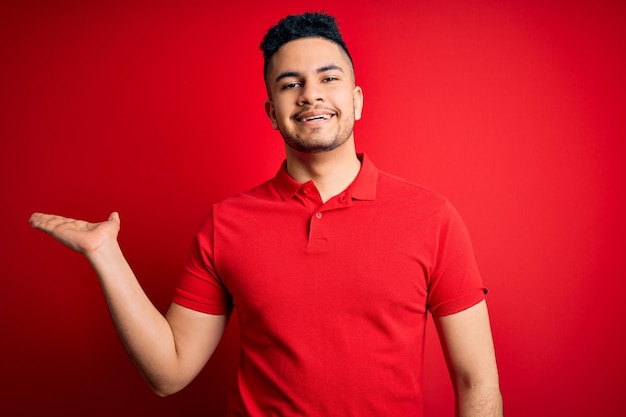 Jonge knappe man met een rode casual polo die over een geïsoleerde achtergrond staat en vrolijk glimlacht en met de handpalm naar de camera wijst