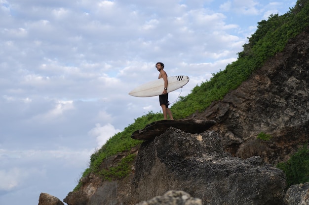 Jonge knappe man met een surfplank op een rots in de buurt van de oceaan.