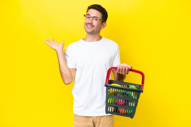 Jonge knappe man met een winkelmandje vol eten over een geïsoleerde gele achtergrond die de handen naar de zijkant uitstrekt om uit te nodigen om te komen