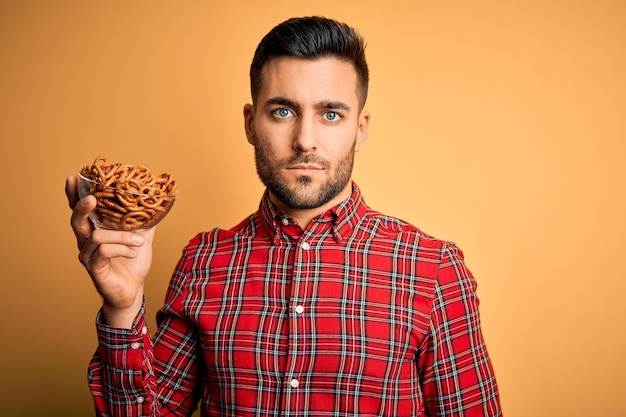 Jonge knappe man met kom met gebakken Duitse pretzels op gele achtergrond met een zelfverzekerde uitdrukking op een slim gezicht die serieus denkt