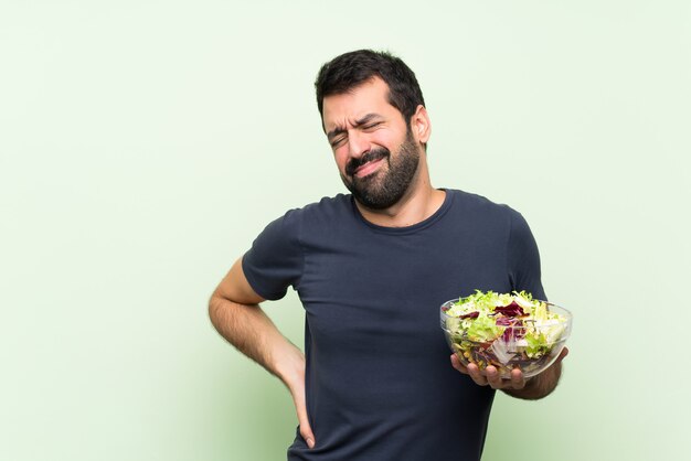 Jonge knappe man met salade over geïsoleerde groene muur die aan rugpijn lijdt omdat hij zich heeft ingespannen