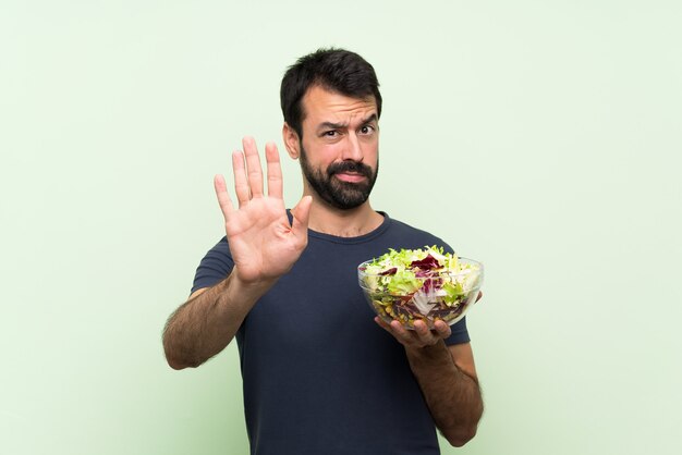 Jonge knappe man met salade over geïsoleerde groene muur stop gebaar maken