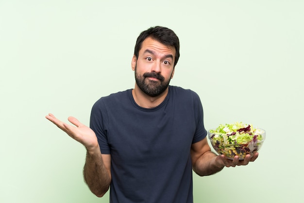 Jonge knappe man met salade over geïsoleerde groene muur twijfels gebaar maken