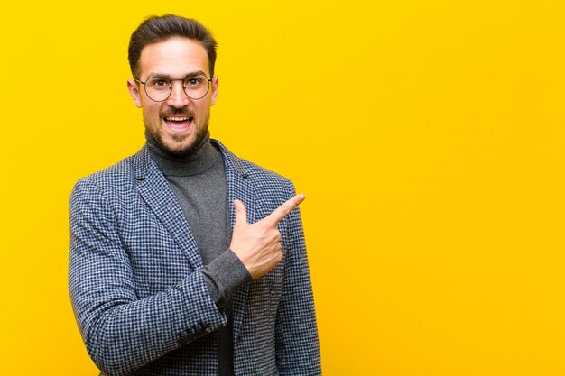 Jonge knappe man op zoek opgewonden en verrast wijzend naar de zijkant en naar boven om ruimte tegen oranje muur te kopiëren