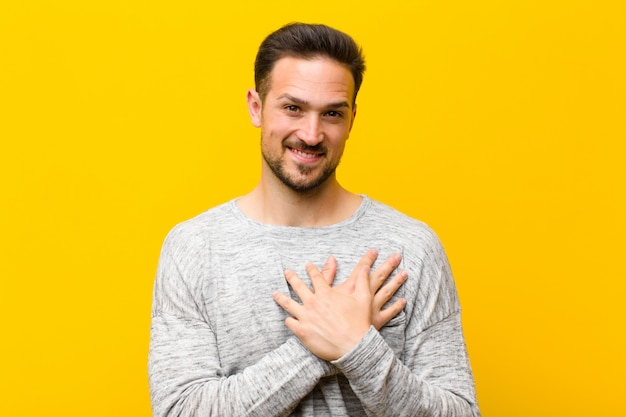 Jonge knappe man romantisch, gelukkig en verliefd voelen, vrolijk glimlachen en hand in hand dicht bij hart tegen oranje muur