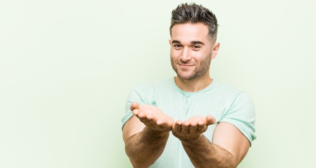 Jonge knappe man tegen een groene achtergrond houden iets met palmen, aanbieden aan camera.