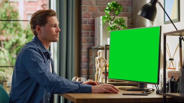 Jonge knappe man werkt thuis op een desktopcomputer met groen scherm Mock Up Display Man controleert bedrijfsrekeningen Boodschappen Collega's Loft woonkamer met groot raam