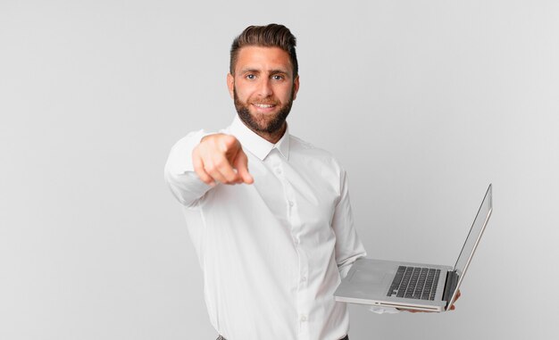 Jonge knappe man wijzend op de camera die jou kiest en een laptop vasthoudt