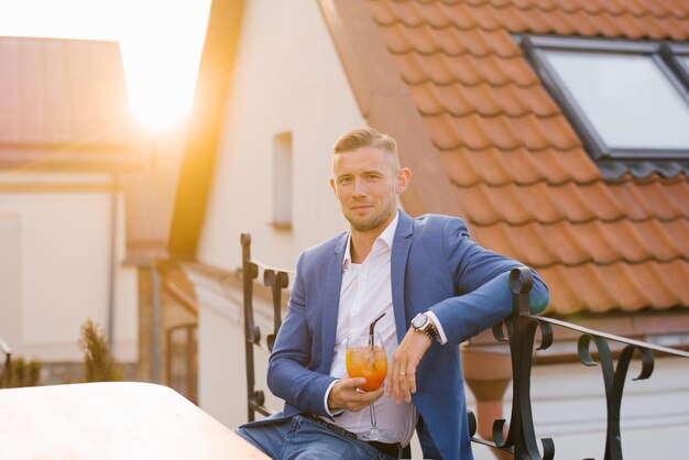 Jonge knappe man zit in een buiten zomer café in een blauw pak en houdt een cocktail glas in zijn hand