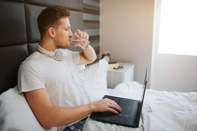 Jonge knappe man zit vanmorgen in bed. hij drinkt water. mannelijke modelgreeplaptop. hoofdtelefoon rond de nek.