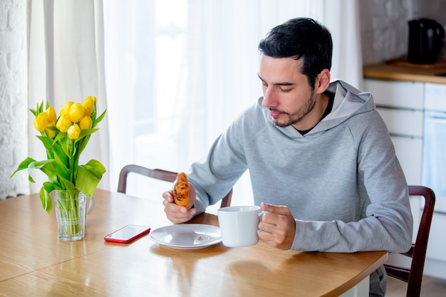 Jonge knappe man zittend aan tafel met een kopje koffie of thee en een croissant. keuken locatie.