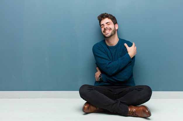 Jonge knappe man zittend op de vloer in liefde voelen, glimlachen, knuffelen en zichzelf knuffelen, single blijven, egoïstisch en egocentrisch zijn