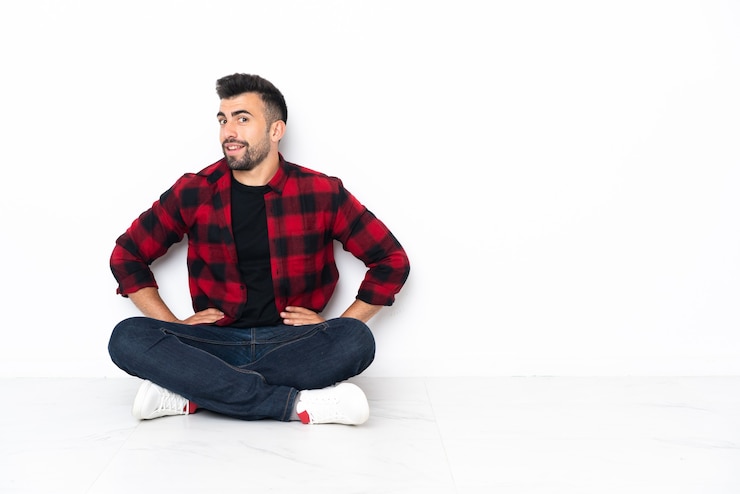 Foto jonge knappe man zittend op de vloer poseren met armen op heup en glimlachend