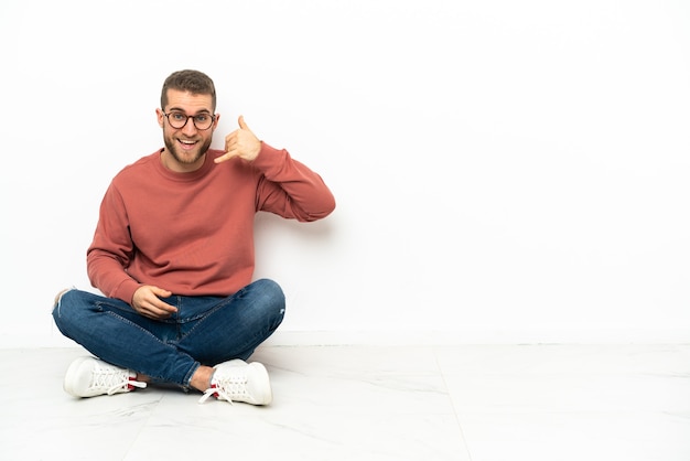 Jonge knappe man zittend op de vloer telefoon gebaar maken. Bel me terug teken