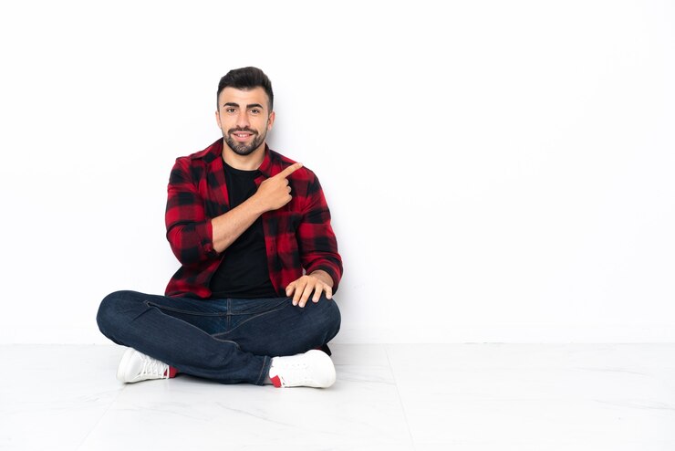 Foto jonge knappe man zittend op de vloer wijzend naar de zijkant om een product te presenteren