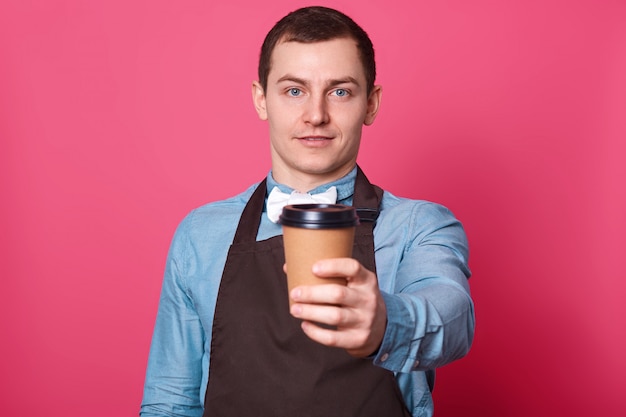 Jonge knappe mannelijke barista stelt voor dat je een kopje koffie van hem maakt, een witte vlinderdas en een bruin schort