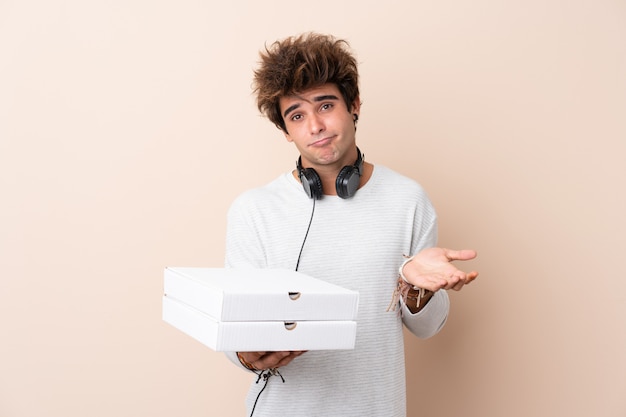 Jonge knappe mens die een pizza over geïsoleerde muur houden die twijfelgebaar maken terwijl het opheffen van de schouders