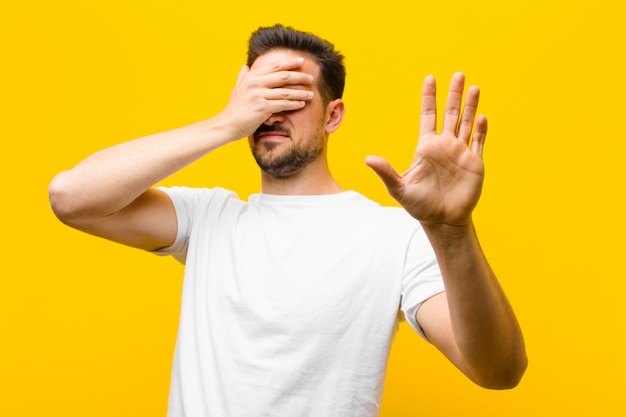 Jonge knappe mens die gezicht behandelen met hand en andere hand vooraan zetten om tegen te houden, weigerend foto's of beelden tegen oranje muur