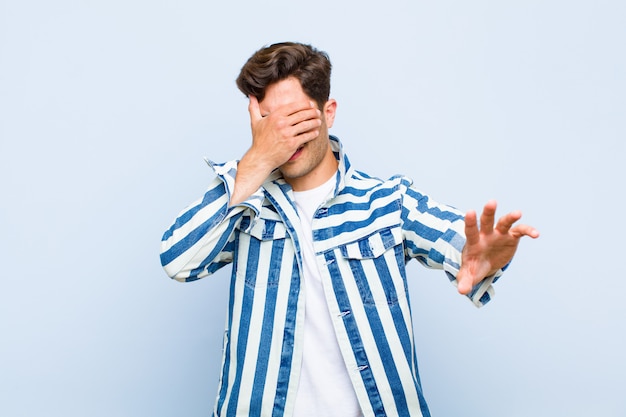 Jonge knappe mens die gezicht behandelt met hand en andere hand vooraan zet om camera tegen te houden, die foto's of beelden over blauwe muur weigert