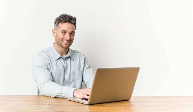 Jonge knappe mens die met zijn gelukkig, glimlachende en vrolijke laptop werkt.