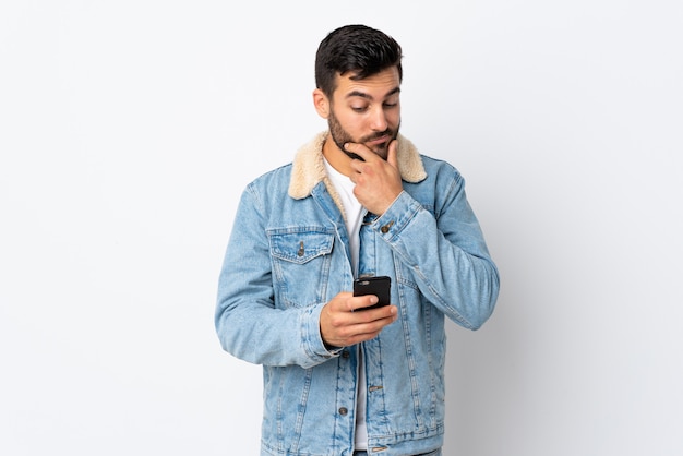 Jonge knappe mens met baard die op witte muur wordt geïsoleerd die en een bericht denkt verzendt