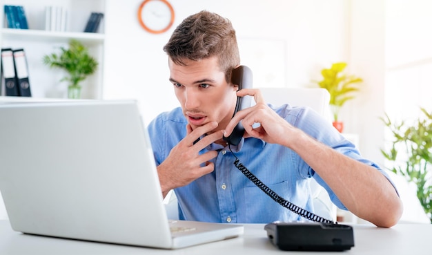 Jonge knappe peinzende zakenman die telefoon gebruikt en naar laptop op kantoor kijkt.